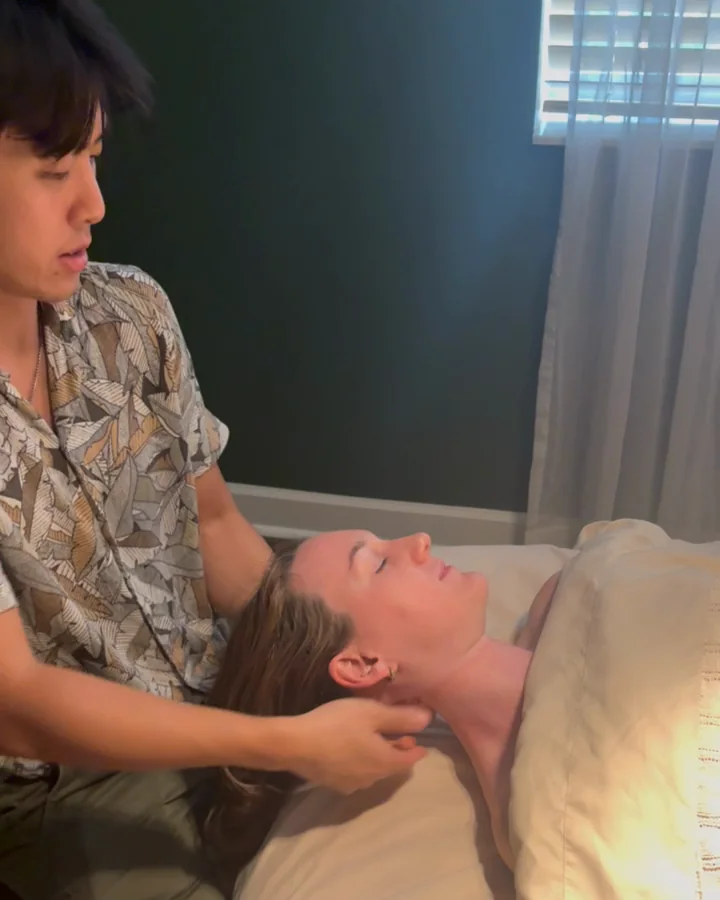 Huan working on the neck of a client who lies face up, draped with a sheet